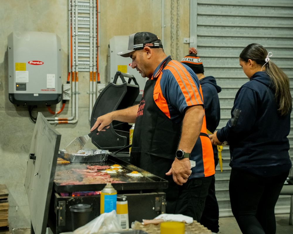 Food service worker in orange safety vest grilling food outdoors near industrial equipment.