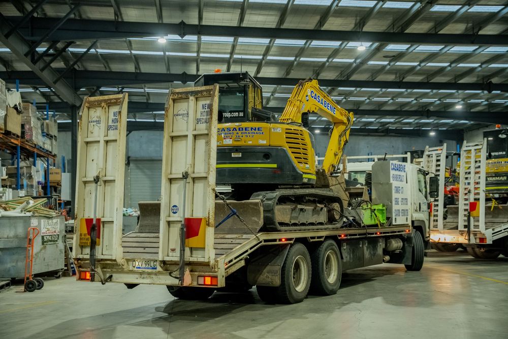 Yellow excavator machine being transported on a flatbed trailer truck inside a warehouse facility.