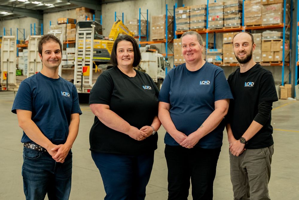 Four warehouse staff members wearing matching navy blue polo shirts stand together smiling in a large storage facility.