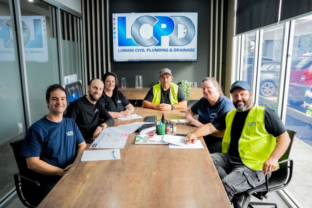 Construction workers have a business meeting around a wooden table with LCPD digital sign visible on wall in background.
