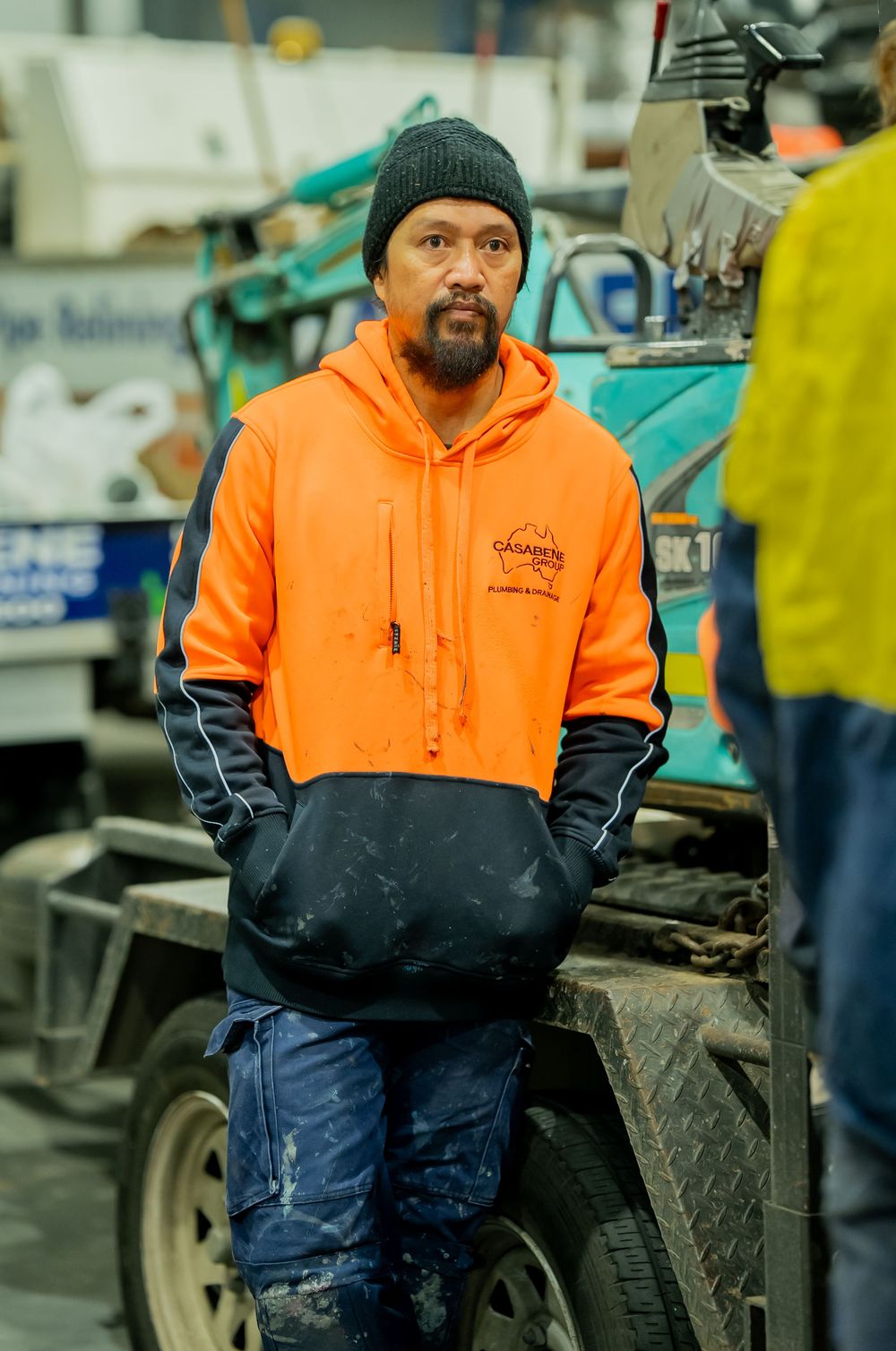 A person wearing an orange and black hooded sweatshirt stands near industrial equipment in a work environment.