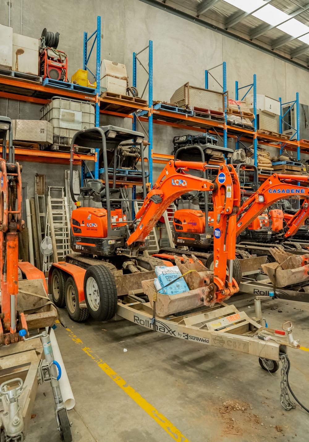 Small orange construction excavators and equipment stored on trailers in an industrial warehouse facility.