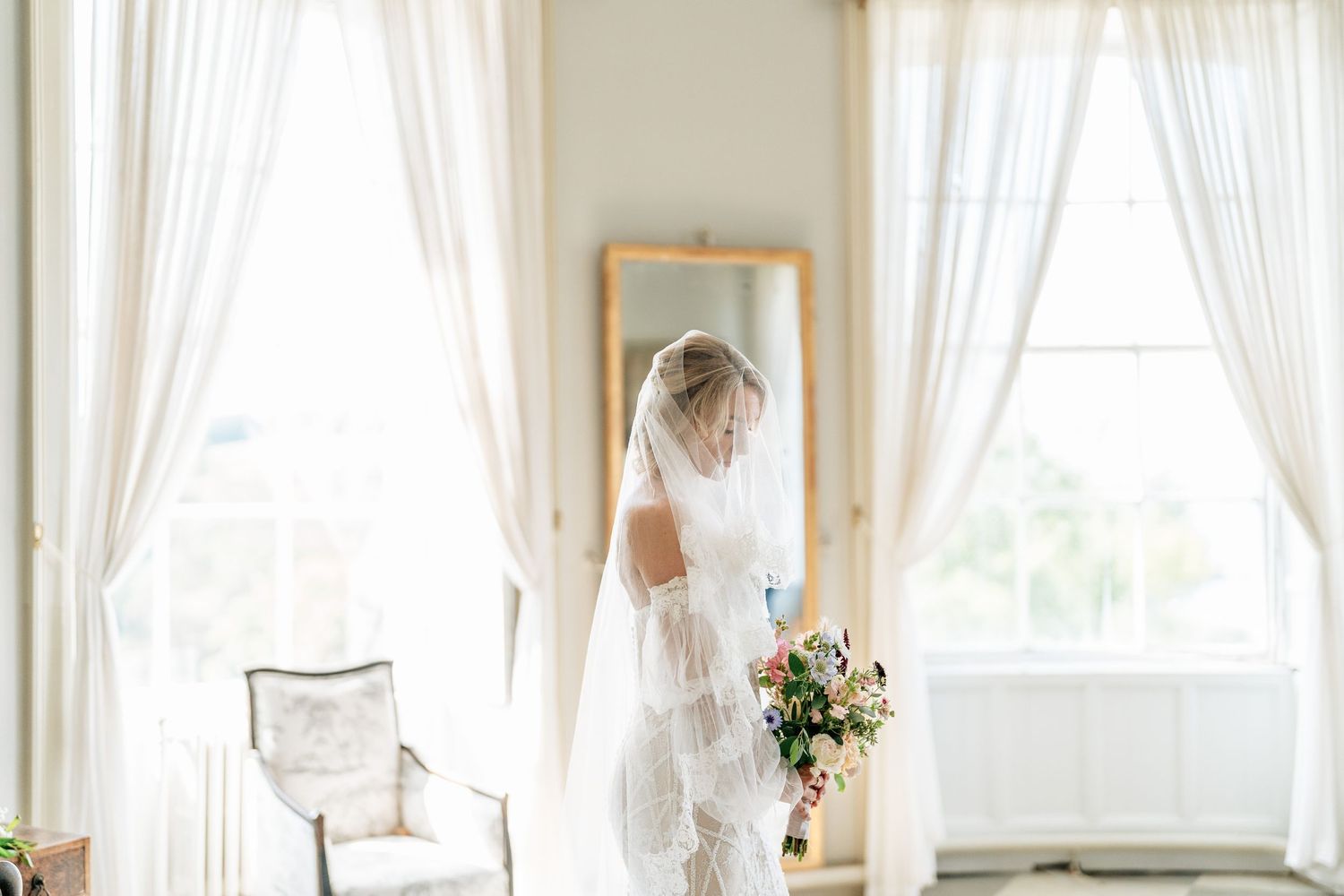 Dreamy bridal reflection in mirror between white curtained windows with soft natural light.