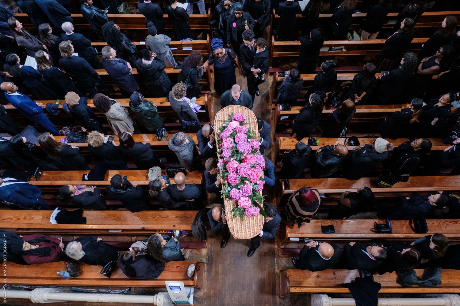 An overhead view of crowd in church pews with pink floral casket in the center during a funeral service.