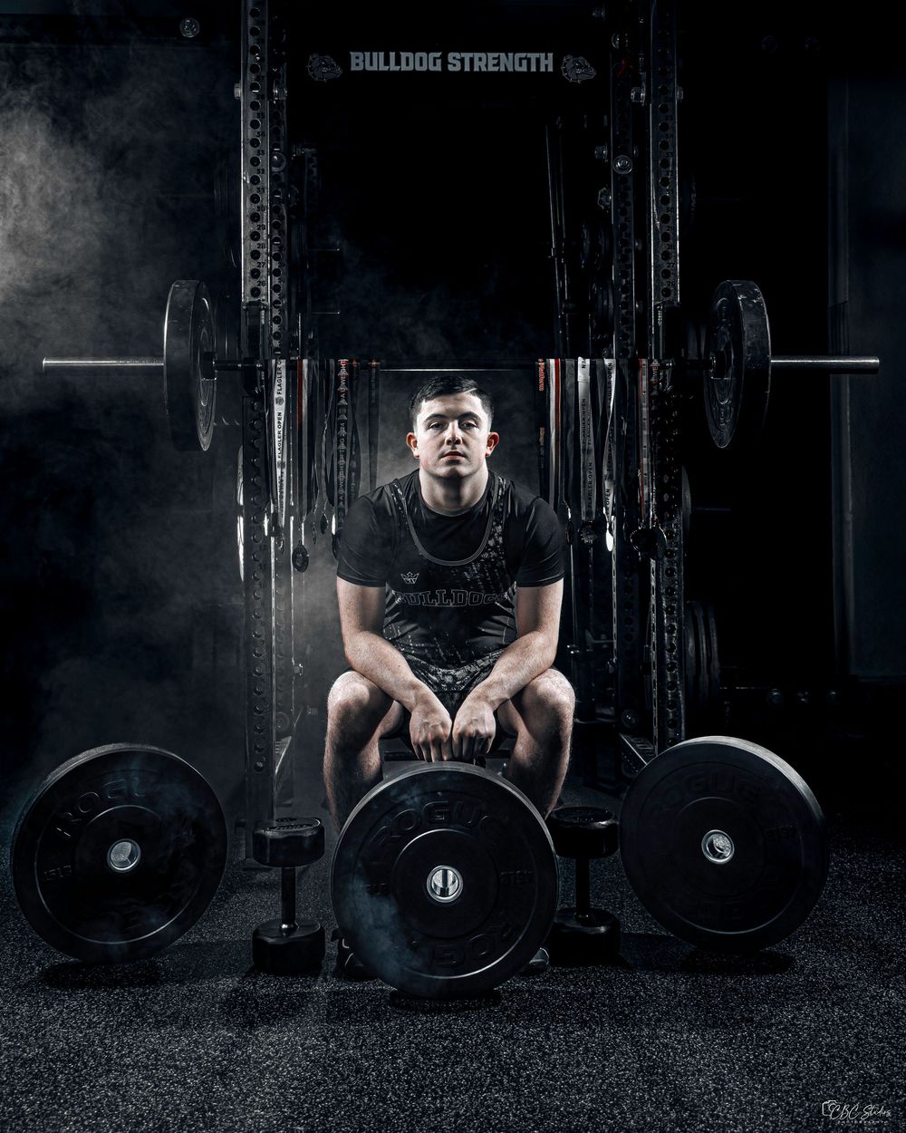 Dramatic gym photo showing a weightlifter resting between sets with heavy barbell plates in dark lighting.