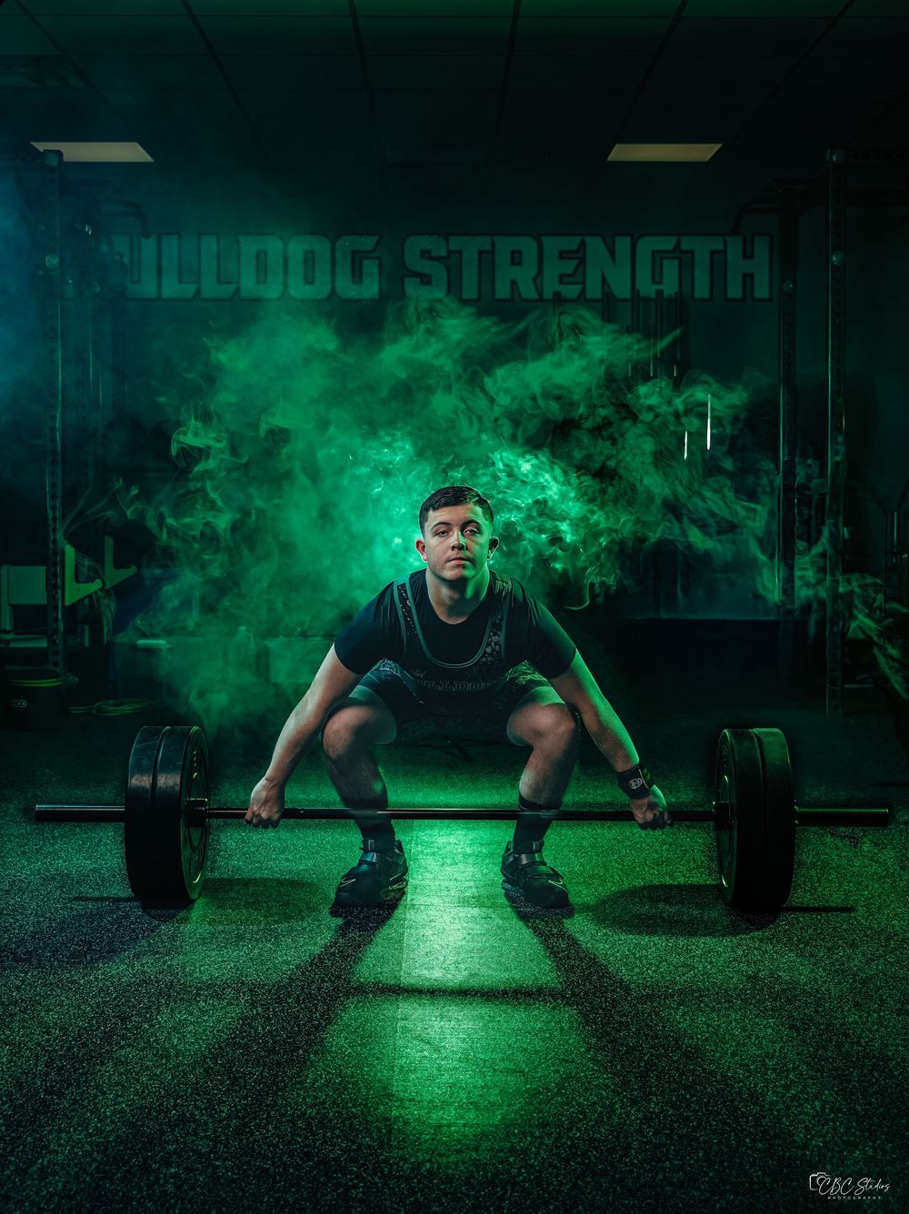 Athletic figure performs deadlift in dark gym with dramatic green smoke effect and 'Bulldog Strength' sign visible.