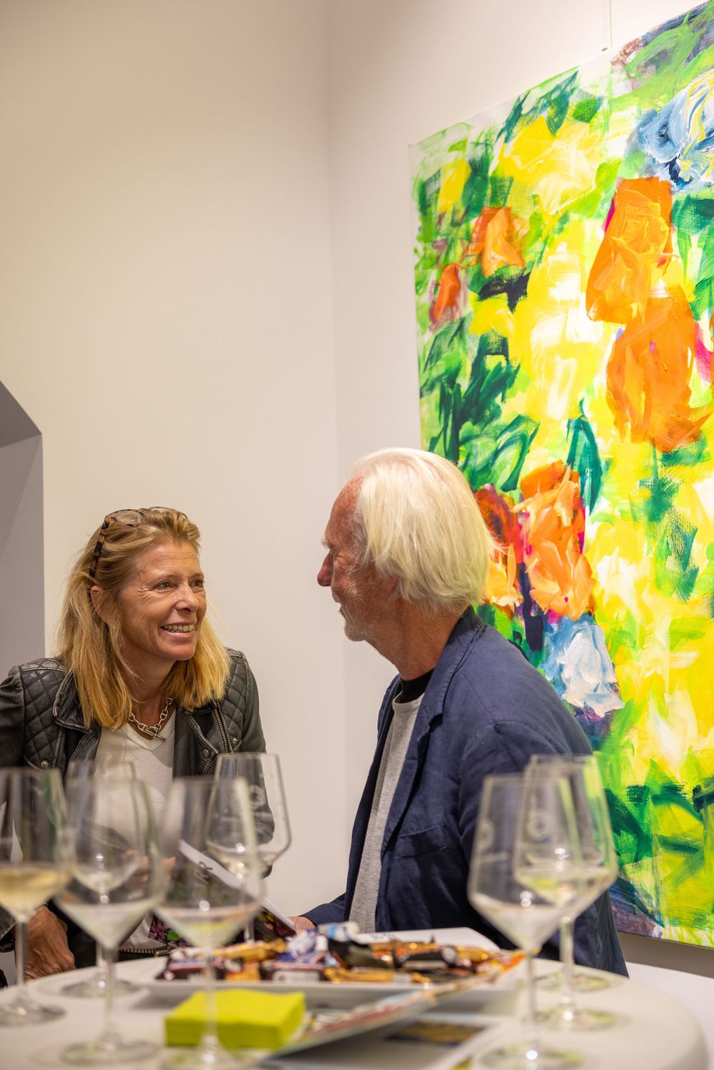 Two people engage in animated conversation at an art gallery while seated near vibrant colorful paintings.