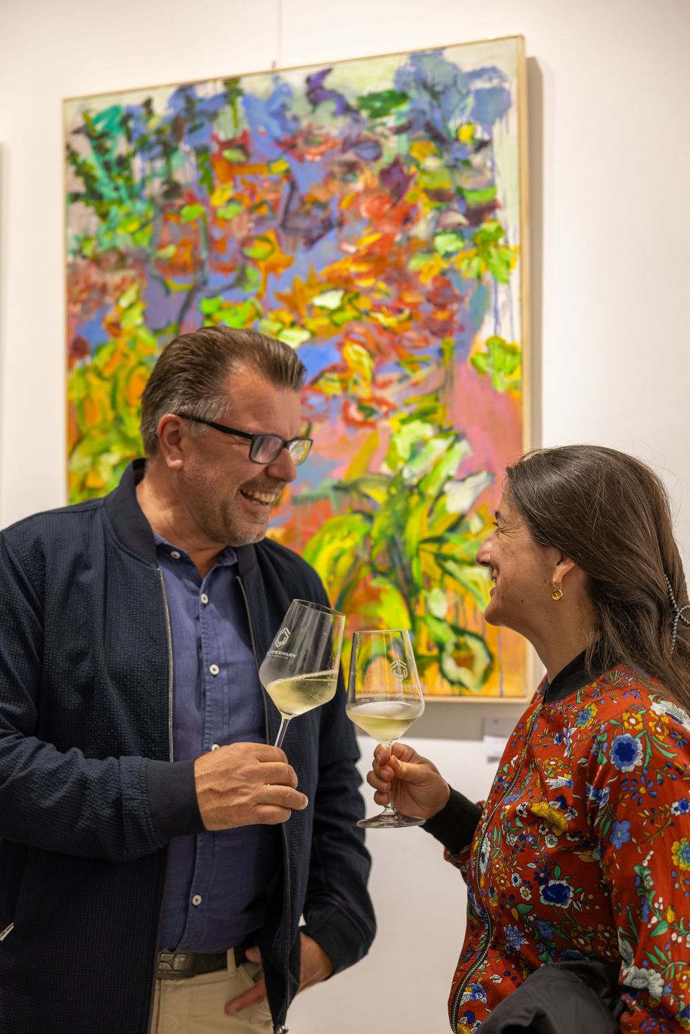 People sharing laughter while viewing vibrant abstract paintings at an art gallery reception.