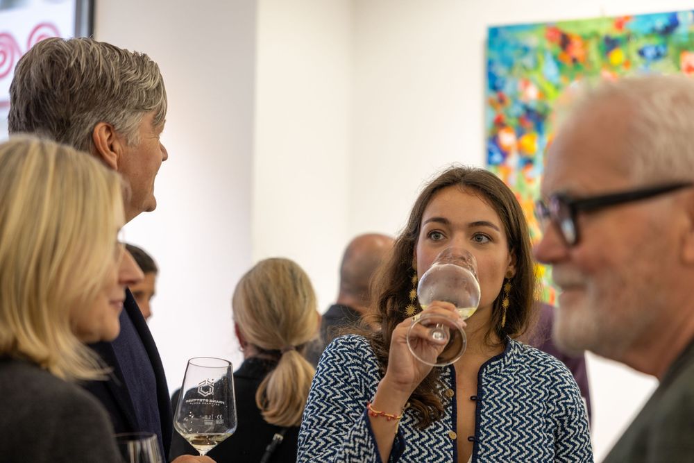 Gallery attendees socialize and laugh together near bright abstract artwork on white walls.