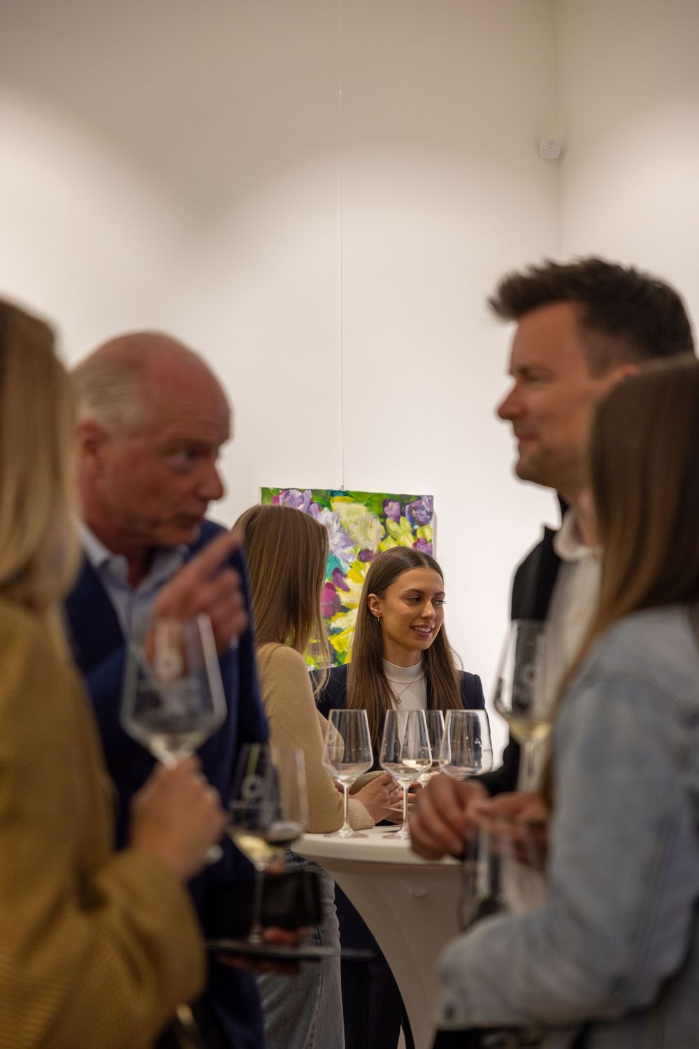 People gather for conversation at an art exhibition with wine glasses and bright paintings.
