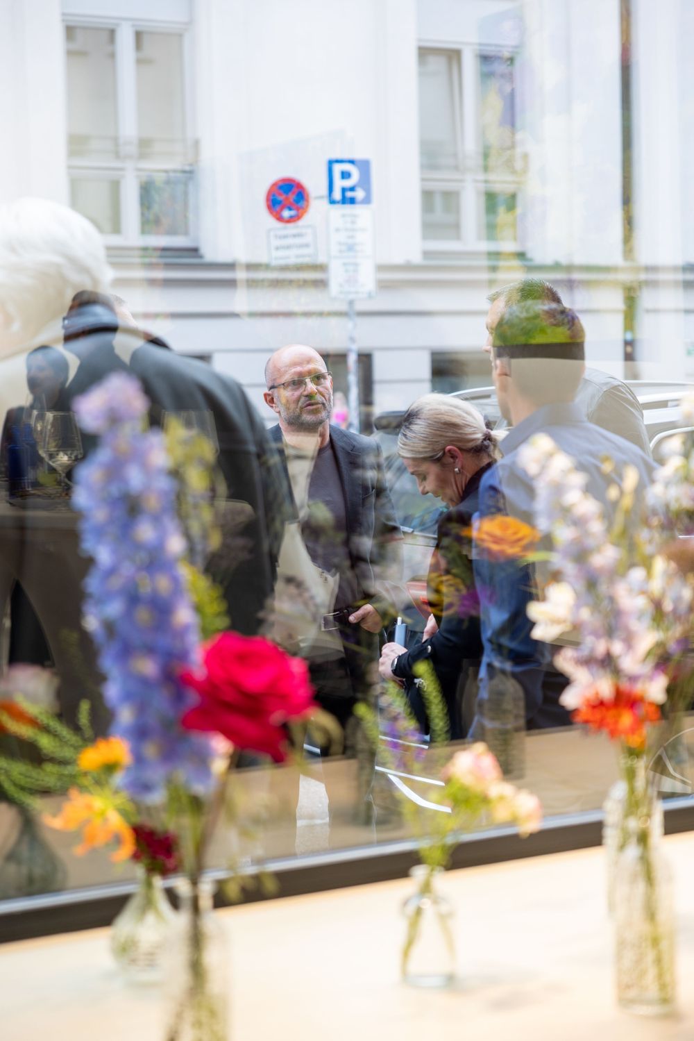 View through window shows gallery visitors with flowers in foreground creating artistic reflections.