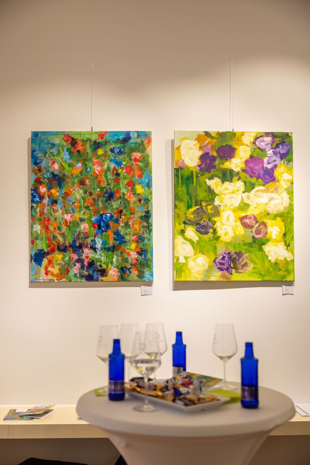 Colorful abstract paintings displayed in series on white gallery wall with glassware on table below.