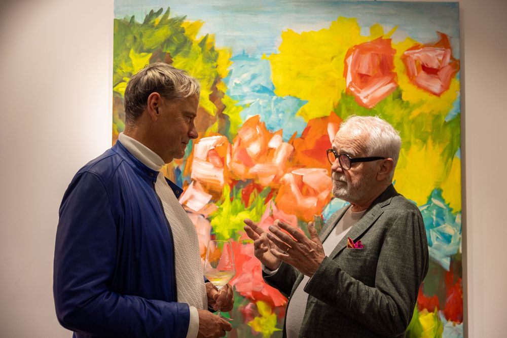 Two attendees discuss artwork in front of bright colorful paintings at gallery opening.