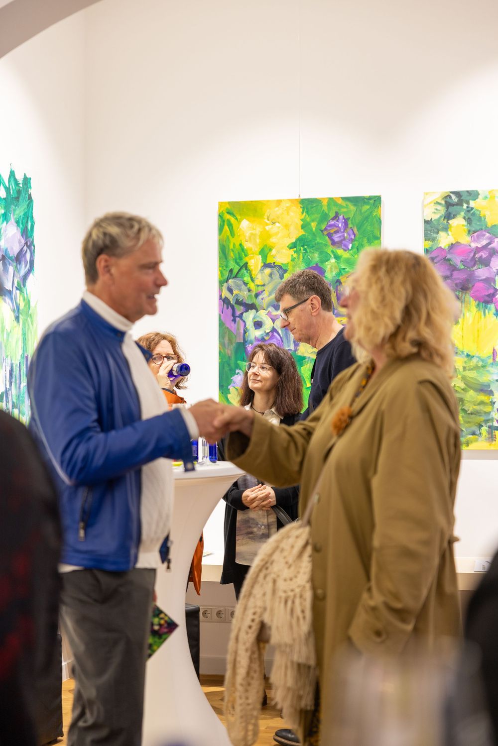 Art gallery attendees interact at an exhibition opening with colorful abstract paintings on display.