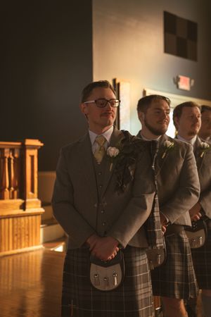 Groomsmen in matching gray suits and kilts standing in line at an indoor venue.