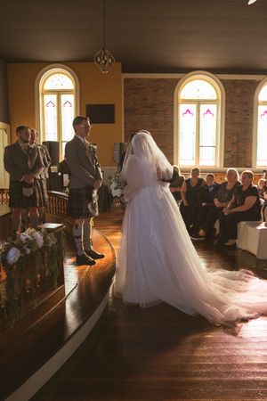 Bride in white gown walks down church aisle during wedding ceremony with sunlight streaming through stained glass windows.