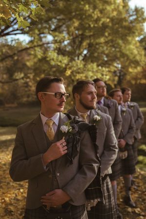 Groomsmen in grey suits and kilts pose together in a fall outdoor setting.