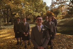Wedding party members stand together on fallen autumn leaves under golden sunlight.