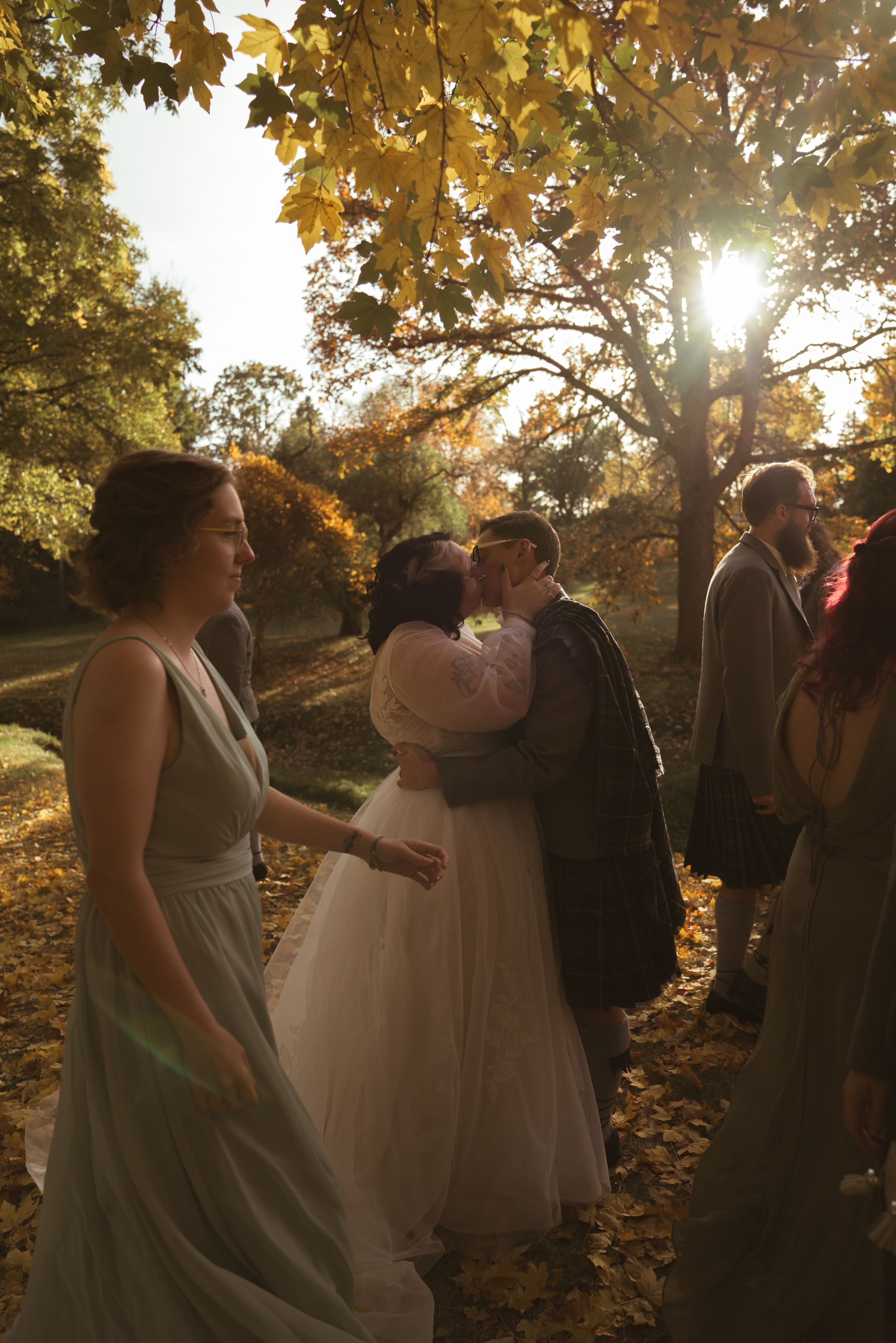 Wedding group gathered under fall trees with golden sunlight filtering through the branches.