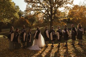 Wedding party poses for photos in a line during golden hour with fall foliage in background.