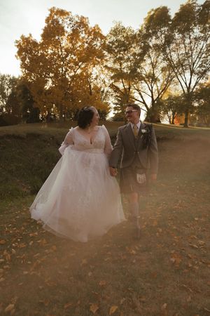 Couple walks hand in hand through fallen leaves during sunset wedding photo session.