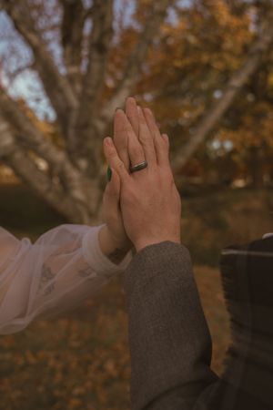 Close-up of hands with wedding rings against blurred fall foliage background.