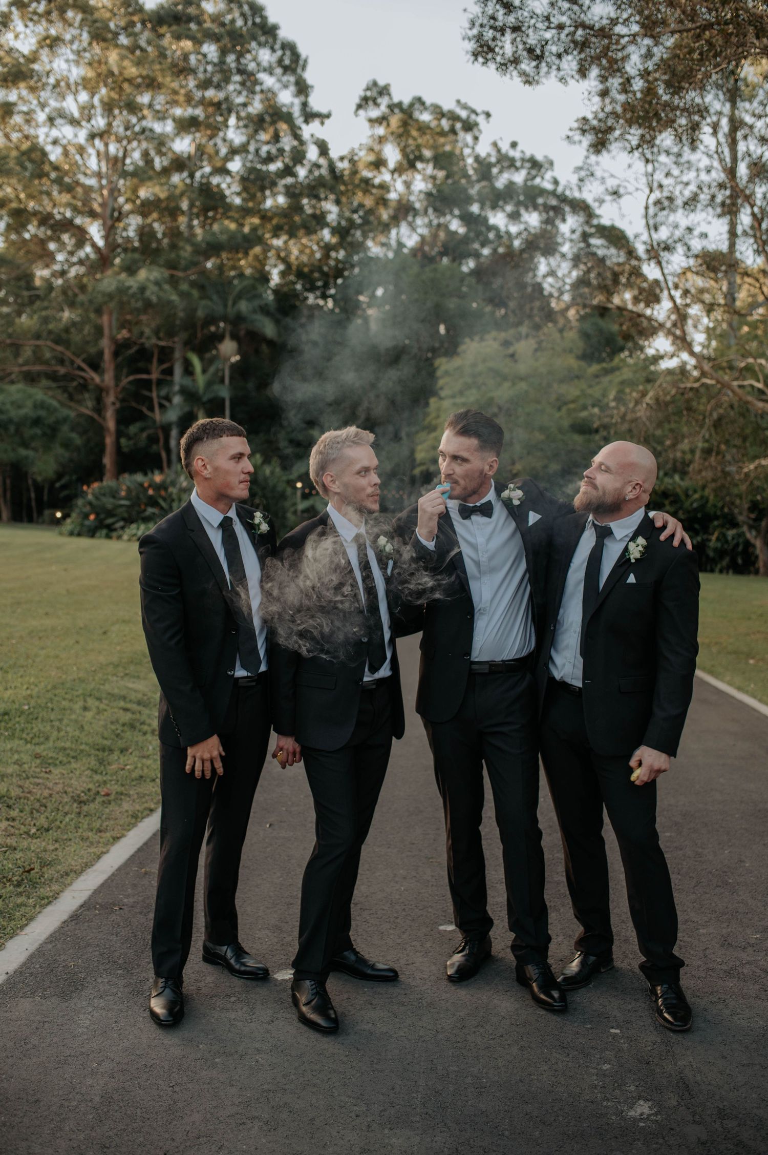 Four groomsmen in black suits share a candid moment laughing together on a path during golden hour.
