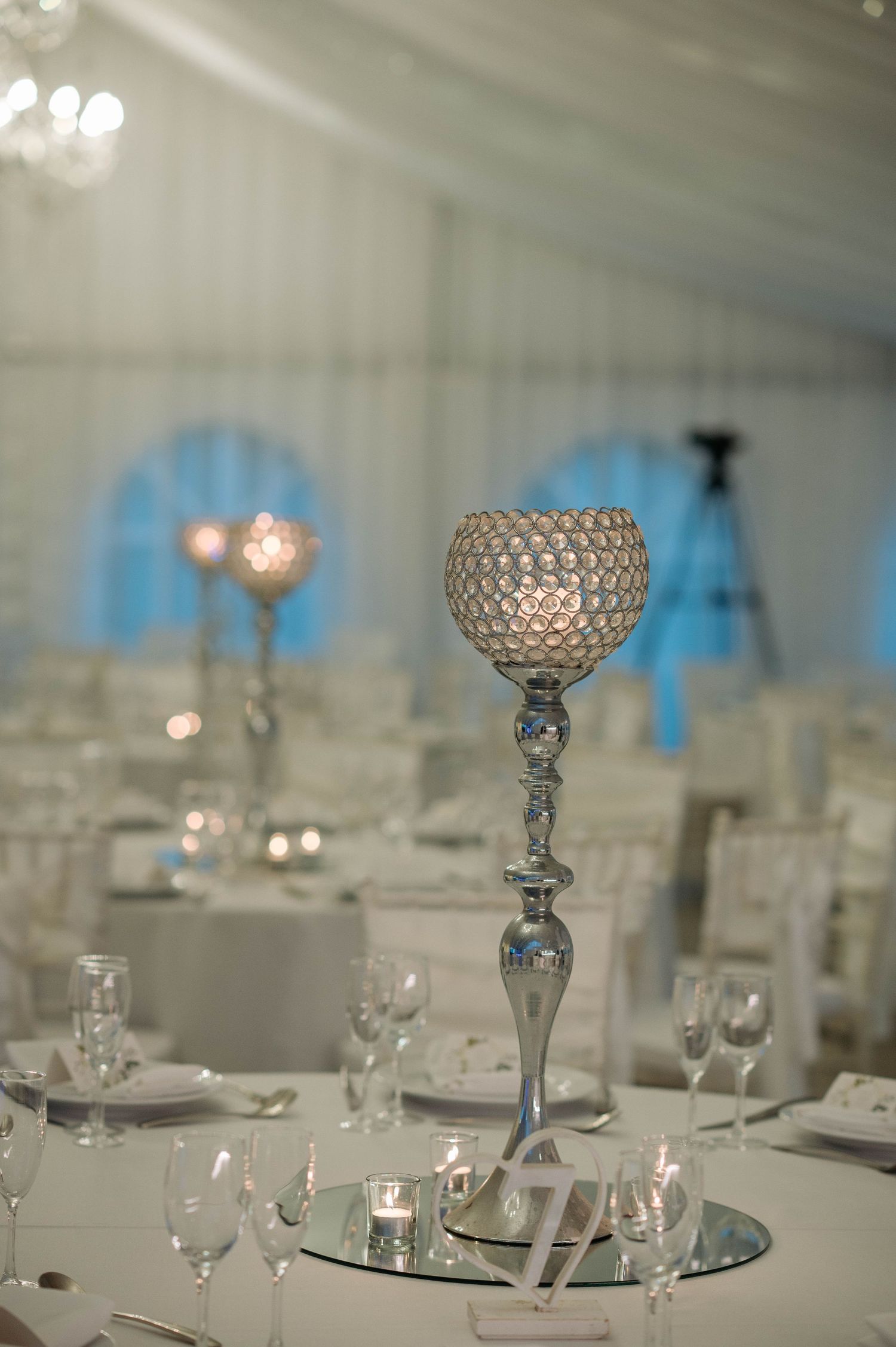 Elegant crystal centerpiece with silver stand and blue backdrop illuminates an upscale wedding reception table setting.
