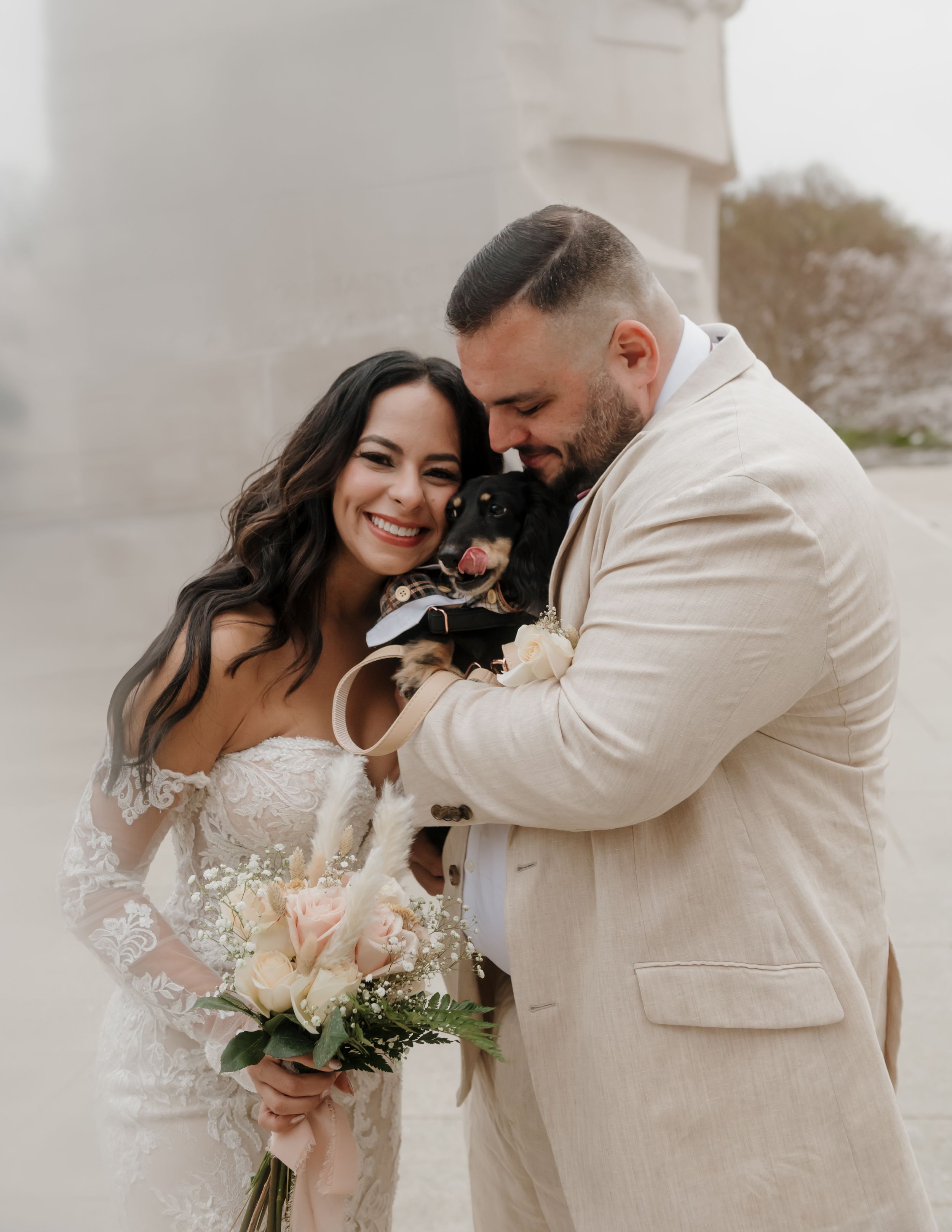 A joyful couple in light-colored wedding attire share a sweet moment with their small dog during their photoshoot.