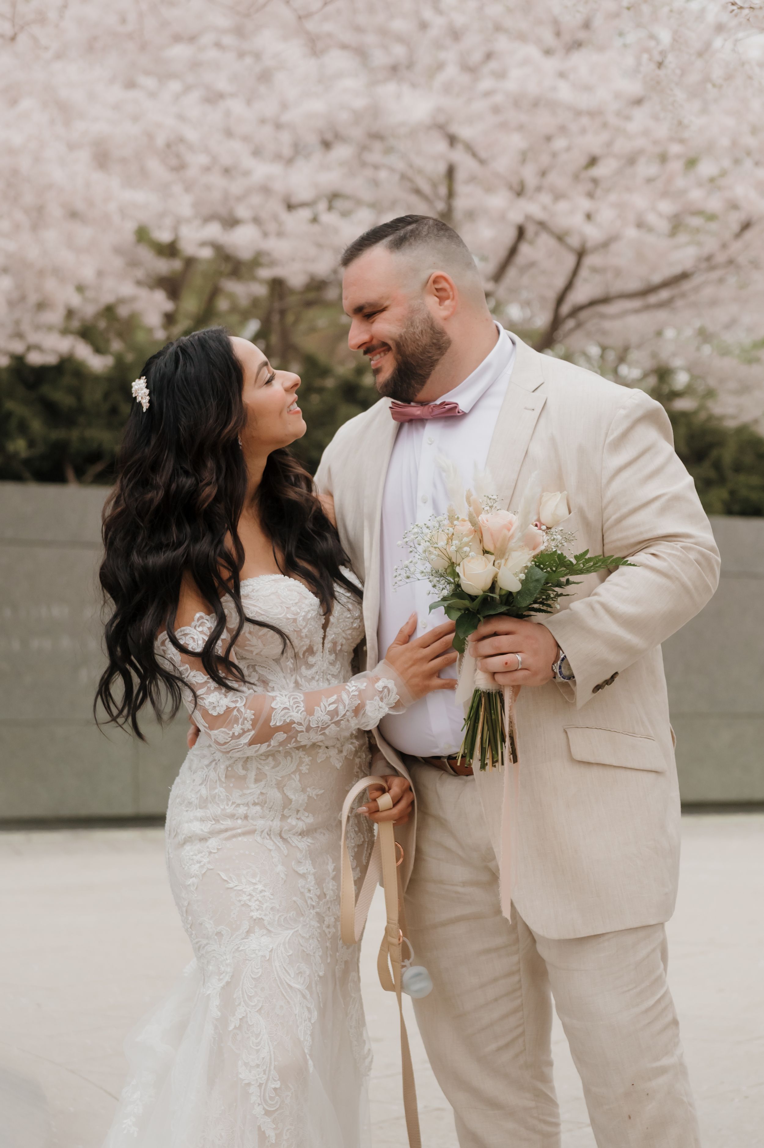 A romantic wedding portrait under cherry blossoms with the couple smiling at each other in cream-colored attire.