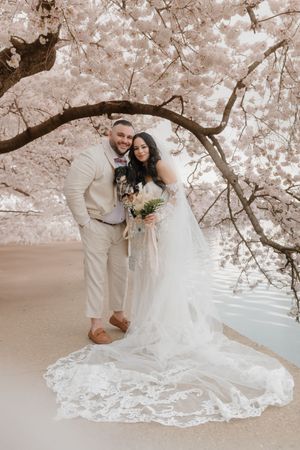 A couple poses under blooming cherry blossoms in springtime while wearing wedding attire near a waterfront.