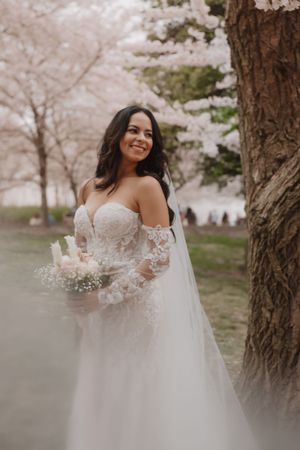 A bride in a lace wedding dress smiles while holding a bouquet under blooming cherry trees.