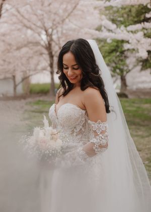 A bride in an off-shoulder lace gown poses with her bouquet under cherry blossoms.