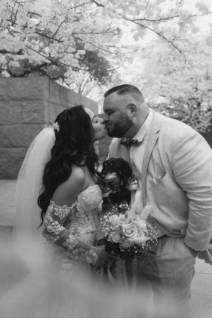 Romantic wedding couple shares intimate moment with their small dog under cherry blossoms in black and white photography.