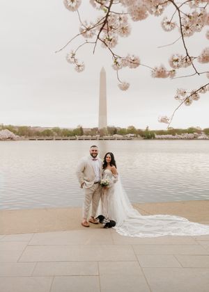 A wedding couple poses by the Washington Monument with cherry blossoms overhead.