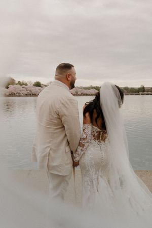 A couple in wedding attire stands by a lake during cherry blossom season in muted tones.
