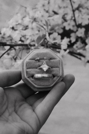 An engagement ring and wedding band nestled in a grey velvet ring box with cherry blossoms in background.