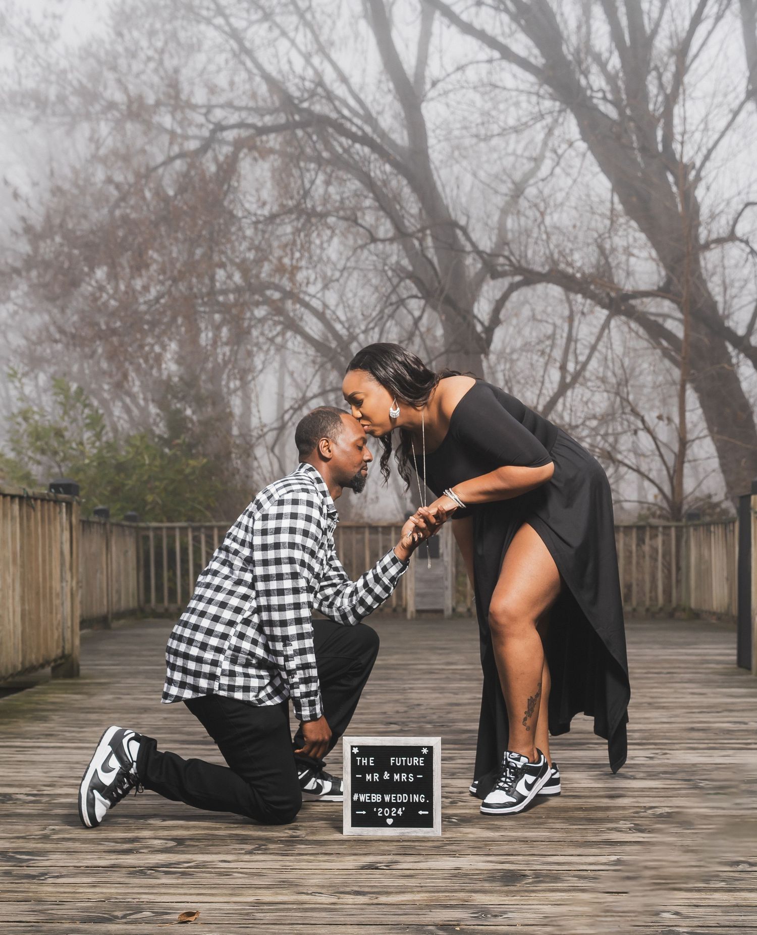 A couple shares a romantic moment on a foggy wooden boardwalk with a small sign board between them.