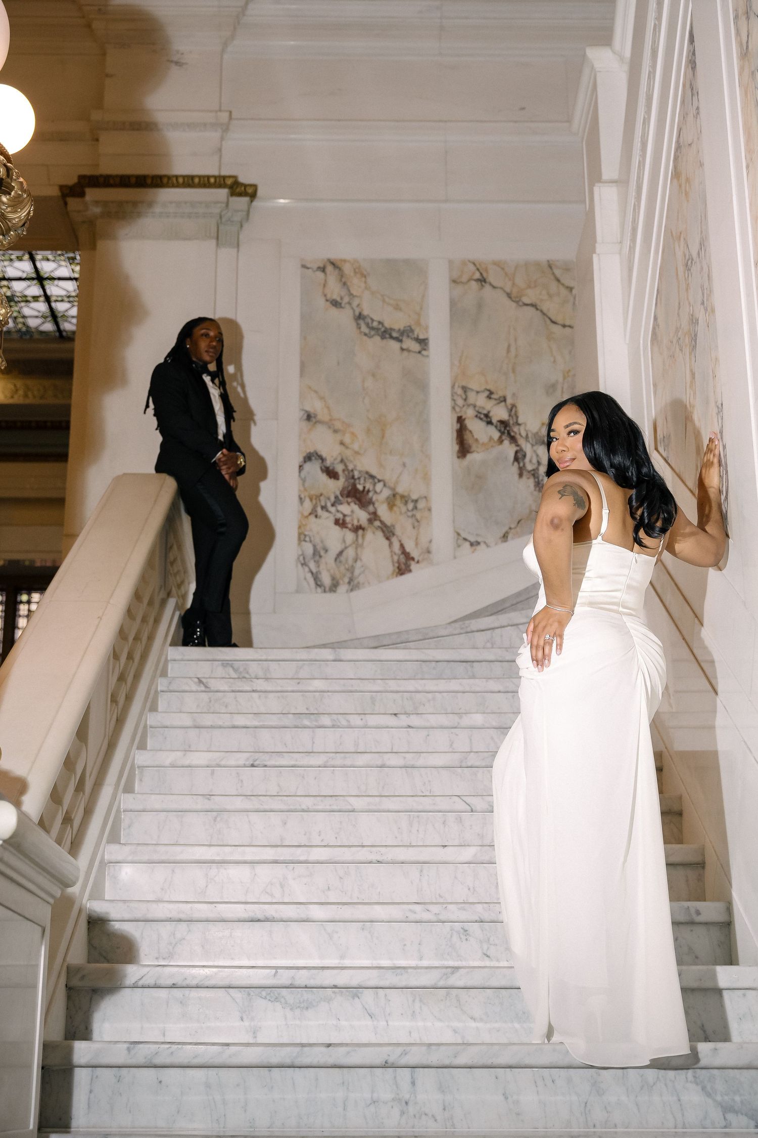 Elegant couple in formal attire posing on marble staircase with decorative walls in grand indoor setting.