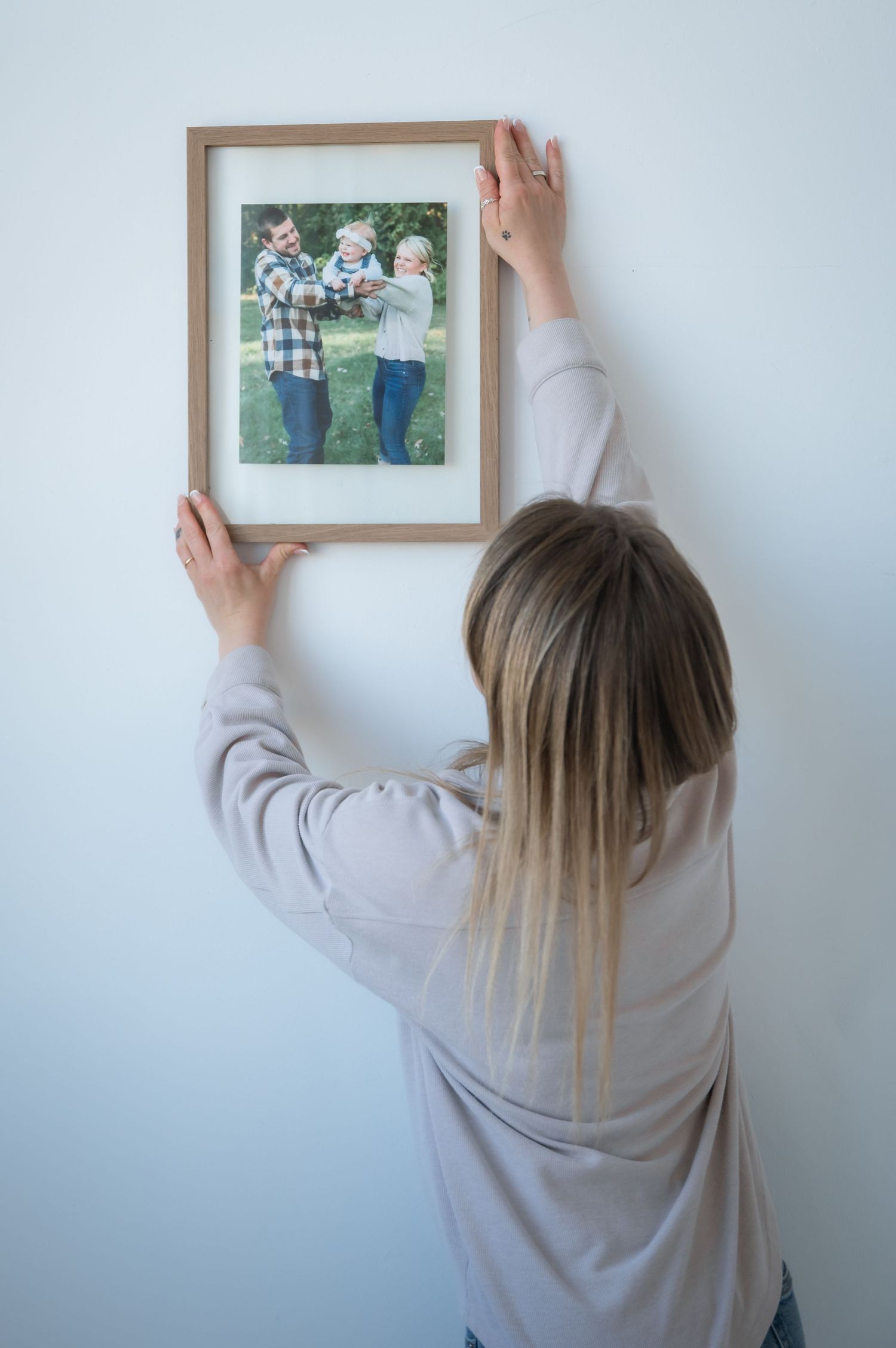 Someone in a gray sweater hangs a wooden picture frame with a photograph on a white wall.