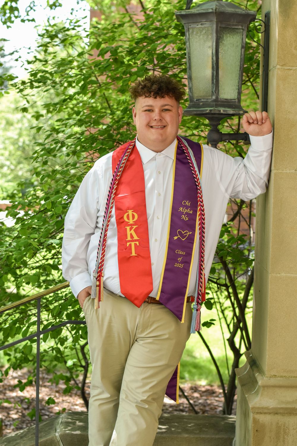 Graduate in khaki pants and red graduation stoles stands by outdoor lamp post during graduation photo session.