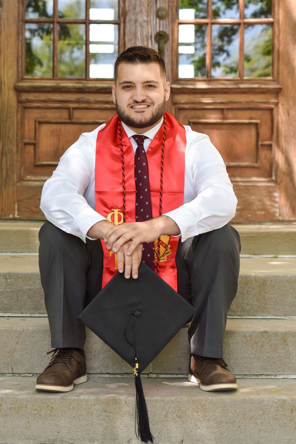 Graduate sits on stone steps wearing red graduation stole, white shirt, and tie with mortarboard cap displayed in front.