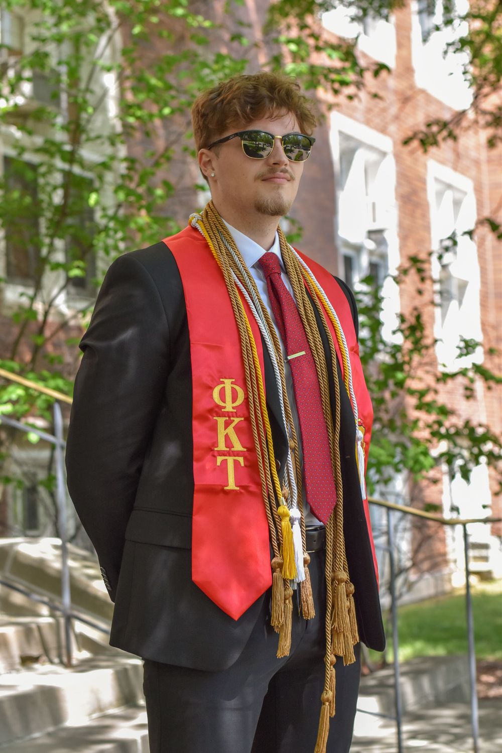 Graduate in black gown and red stola with honor society cords standing outside during commencement ceremony.