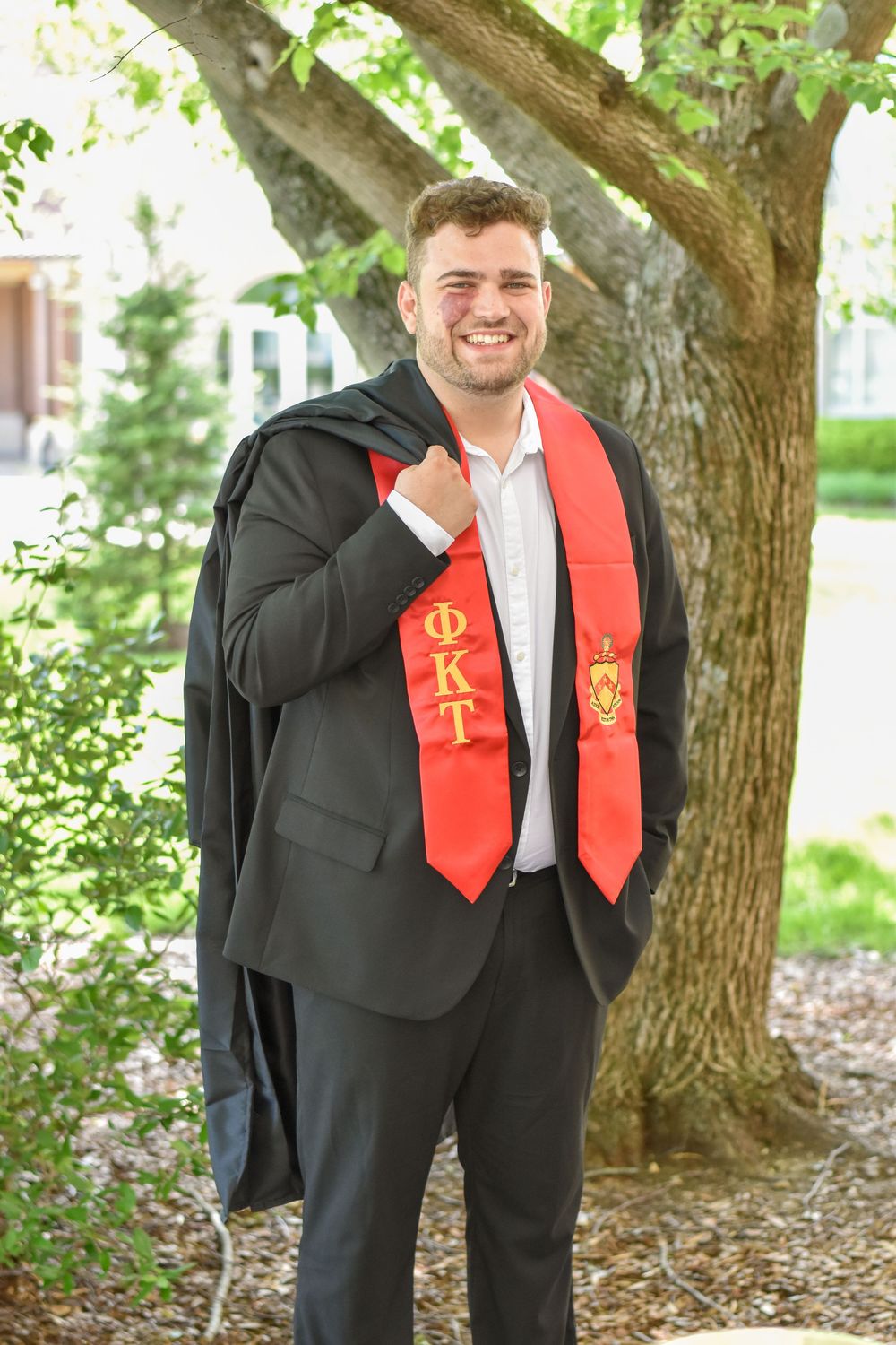 Graduate in dark suit and red graduation stole stands smiling under a tree during commencement.