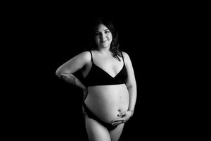 Black and white maternity photo of woman in lingerie holding her pregnant belly against dark background.