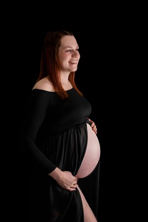 A maternity portrait against black background shows an expectant mother in a long-sleeved black dress smiling warmly.