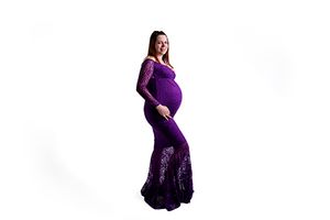 A pregnant woman poses in a long purple maternity gown with lace details against a white background.