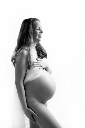 A black and white maternity photo of an expectant mother in profile wearing light clothing against a plain backdrop.