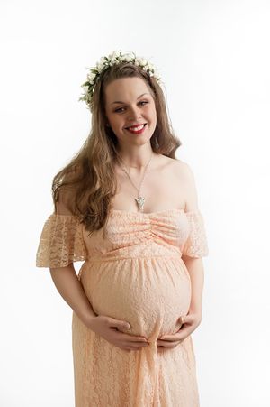 Pregnant woman in peach-colored lace dress and floral crown stands against white background with hands on belly.