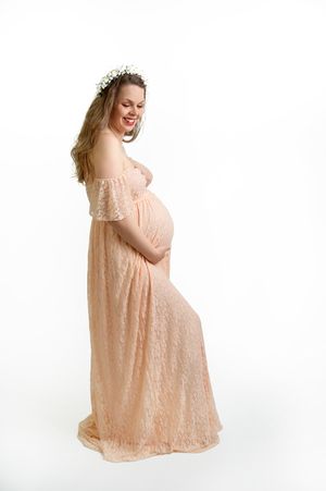 A joyful expectant woman in a peach lace dress and floral headpiece poses against a white backdrop.